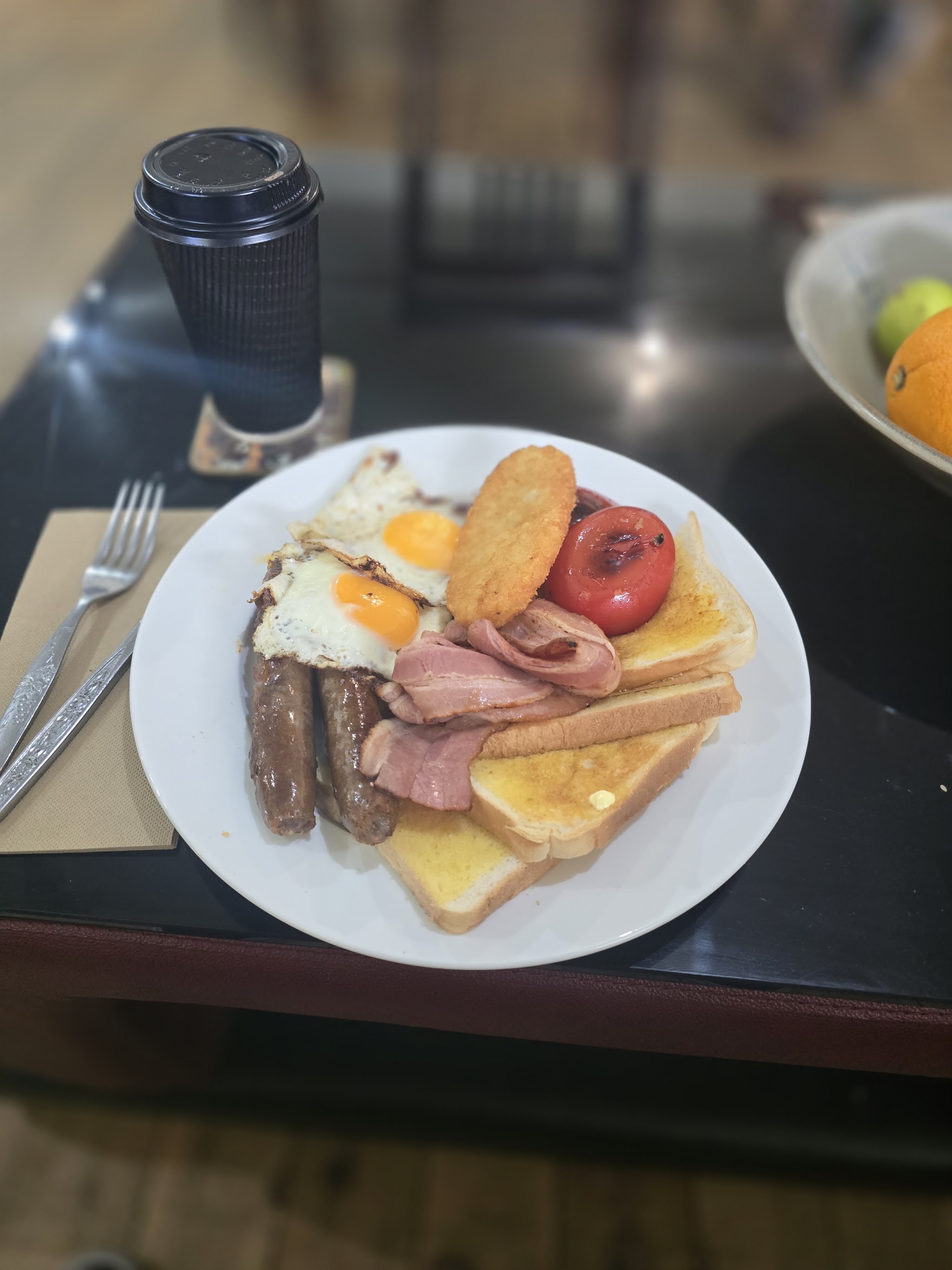 A full cooked plate of eggs, bacon, sausages, hash brown, tomato and toast with a coffee
